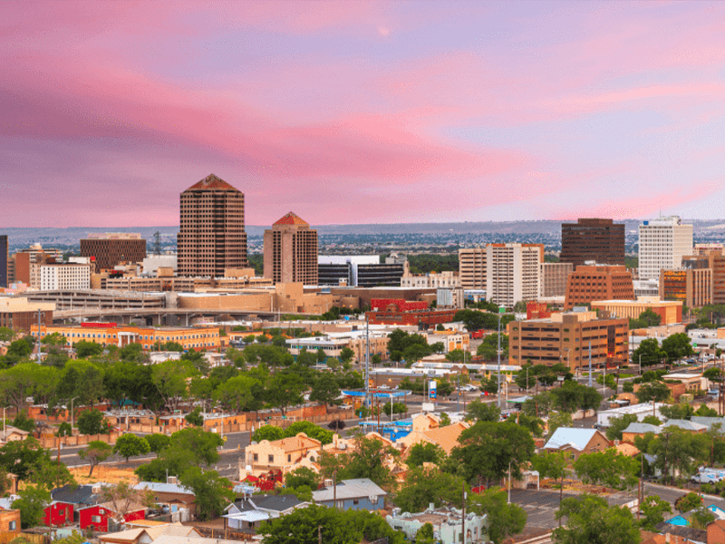 A cityscape view of downtown Albuquerque, New Mexico, with modern buildings, trees, and houses under a pink and purple sunset sky.