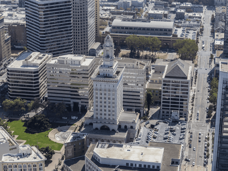 Aerial view of downtown Oakland, California, featuring the historic white Oakland City Hall building with its clock tower, surrounded by modern office buildings, streets, and parked cars.