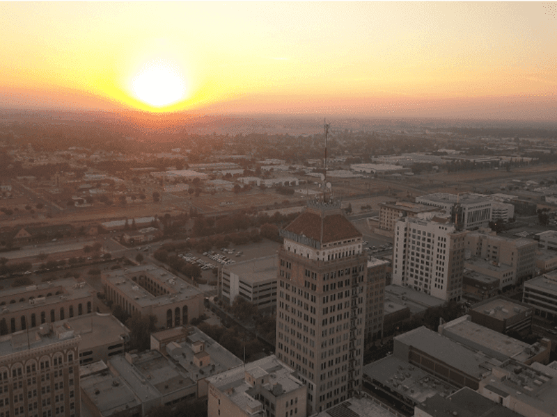 Aerial view of a cityscape at sunset, with the sun low on the horizon, casting a warm glow over buildings, streets, and a tall tower in the foreground.