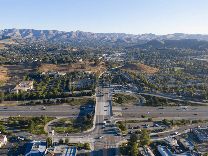 Aerial view of a wide intersection with multiple lanes and cars, surrounded by greenery, buildings, and hills in the distance under a clear blue sky.