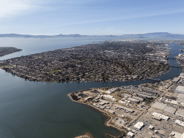 Aerial view of an urban peninsula surrounded by water, with industrial buildings in the foreground, residential neighborhoods in the middle, and mountains and a distant city skyline under a clear sky.