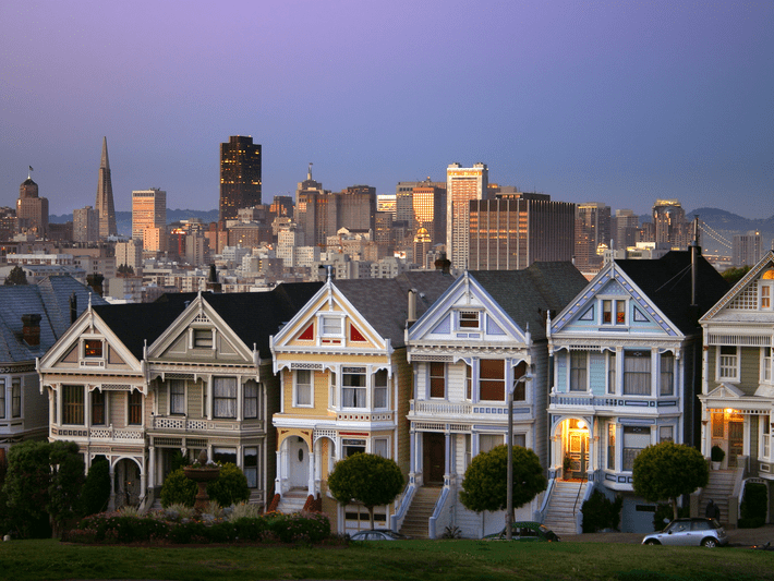 A row of colorful Victorian houses, known as the Painted Ladies, stands in front of the San Francisco skyline at dusk, with city buildings illuminated in the background.