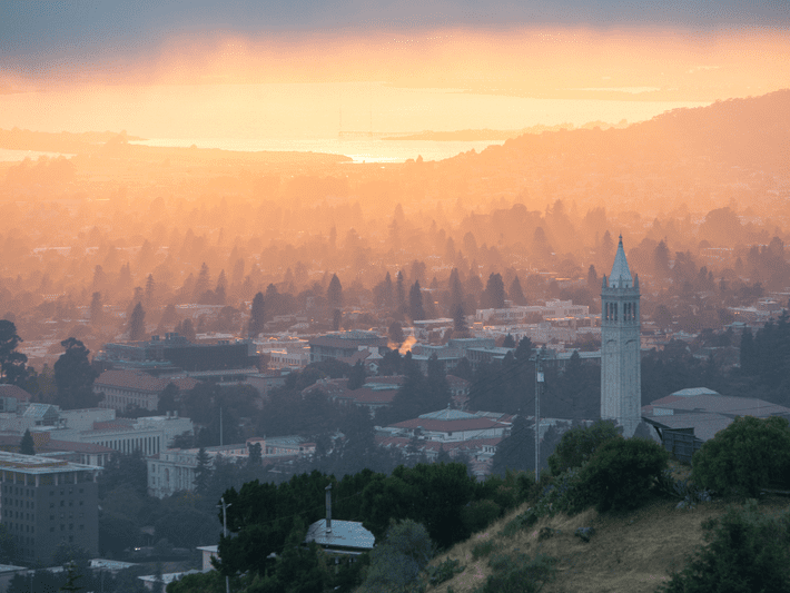 View of a city at sunset with a tall clock tower, soft orange light, and misty atmosphere. Trees and buildings fill the landscape, hills in the foreground, and a bay visible in the distant background.