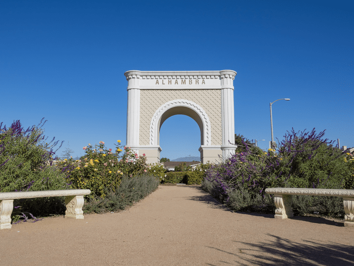 A large white archway labeled “Alhambra” stands at the end of a garden path, surrounded by flowering shrubs, benches, and greenery under a clear blue sky.