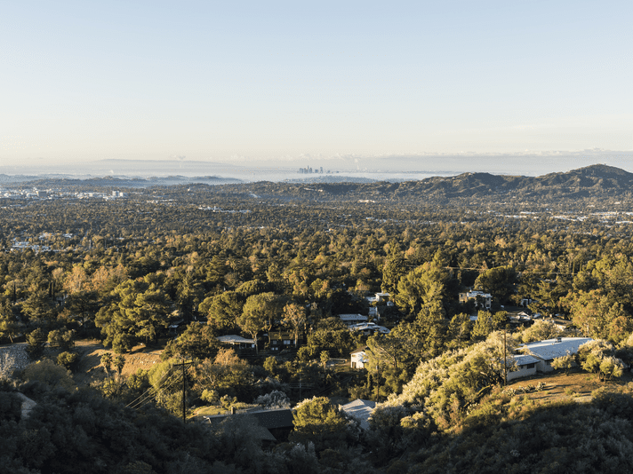 A wide view of a city landscape with scattered houses among dense green trees, rolling hills, and a distant skyline under a clear sky.