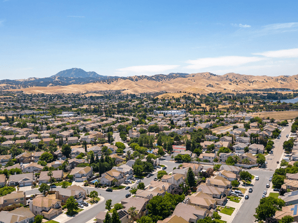 Aerial view of a suburban neighborhood with rows of houses, tree-lined streets, and a background of rolling golden hills and a distant mountain under a clear blue sky.