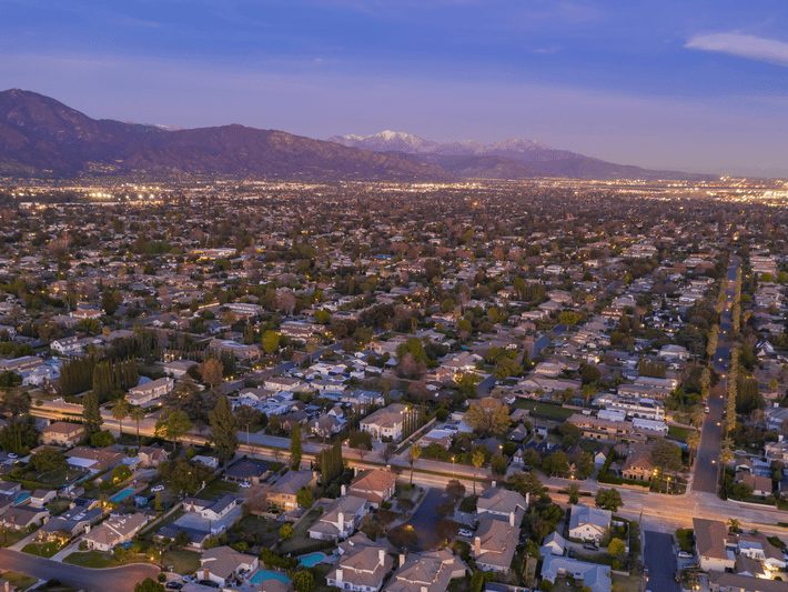 Aerial view of a suburban neighborhood at dusk, with rows of houses, tree-lined streets, and mountains in the background under a partly cloudy sky. City lights begin to illuminate the distant horizon.
