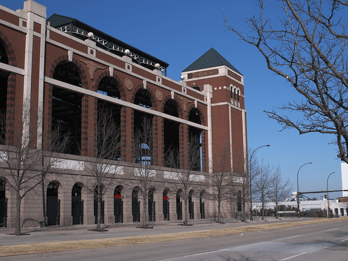 A large, brick stadium with tall arched windows and a tower sits beside an empty street lined with leafless trees under a clear blue sky.