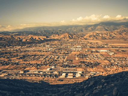 A sepia-toned aerial view of a sprawling town in a dry, desert landscape, surrounded by mountains under a cloudy sky.