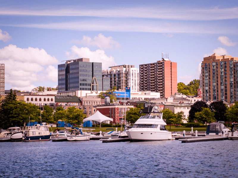 A marina with several boats docked on calm water, trees lining the shore, and mid-rise buildings and apartments in the background under a partly cloudy sky.
