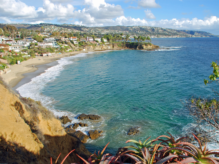 A scenic coastal view of a sandy beach bordered by rocky cliffs and lush green plants, with waves gently reaching the shore. Houses line the hills above the beach under a partly cloudy sky.