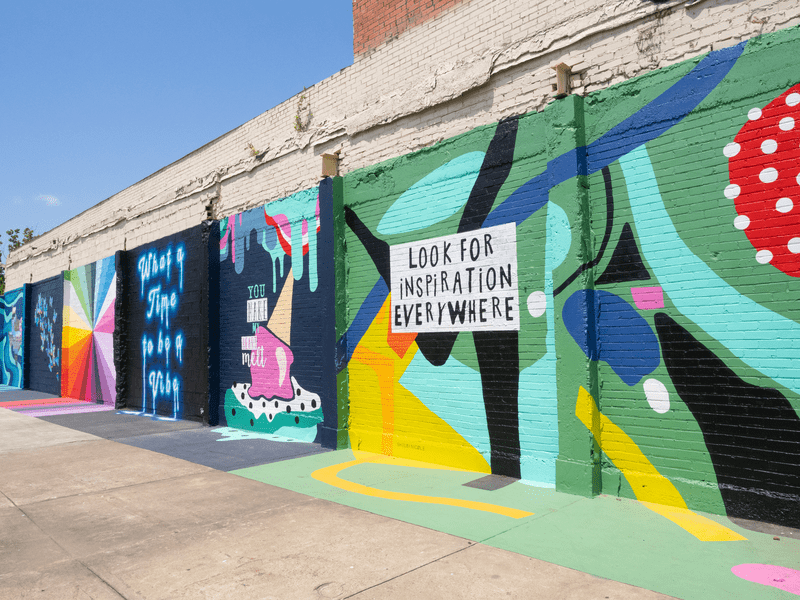 A colorful mural on a brick wall features bold shapes, abstract patterns, and the phrase “LOOK FOR INSPIRATION EVERYWHERE” in black letters on a white background. Sidewalk and blue sky are also visible.