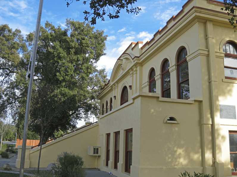 A yellow, historic building with arched windows and red trim stands next to tall trees under a blue sky with scattered clouds.