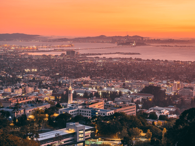 A vibrant cityscape at sunset shows a bay with a bridge, urban buildings, and distant mountains under an orange sky. The foreground has a university campus with a tall clock tower.