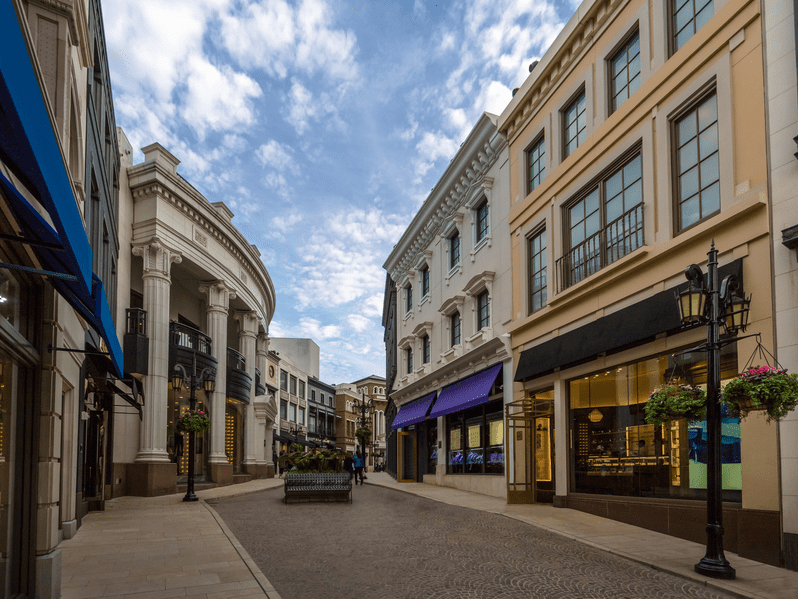 A clean, upscale shopping street with elegant buildings, large windows, and awnings, some in blue and black. The cobblestone path is empty under a partly cloudy sky. Hanging flower baskets decorate lampposts.