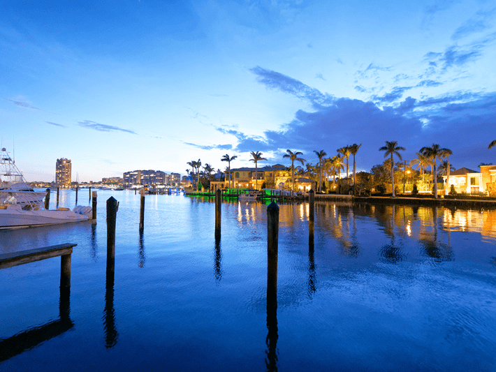 A calm waterfront scene at sunset with palm trees, dock posts reflected in the water, moored boats, and city buildings lit up in the distance under a blue sky.