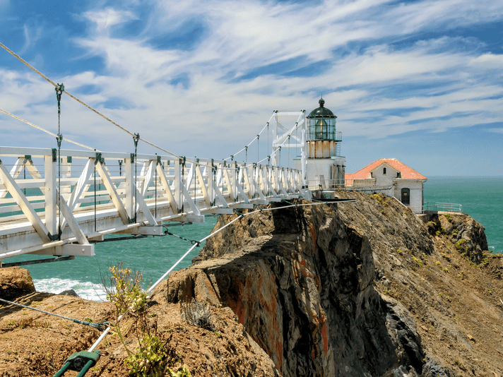 A white suspension bridge leads to a historic lighthouse and small building perched on a rocky cliff above the ocean under a blue sky with wispy clouds.