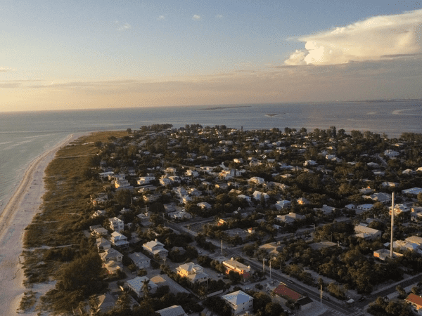 Aerial view of a coastal town at sunset, with houses nestled among trees near a sandy beach and calm ocean waters stretching to the horizon under a partly cloudy sky.