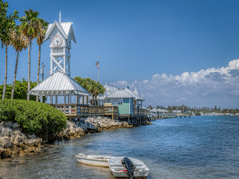 A small boat floats near the rocky shoreline; a white wooden clock tower with a tin roof stands on a pier above the blue water, with palm trees and buildings under a sunny sky in the background.