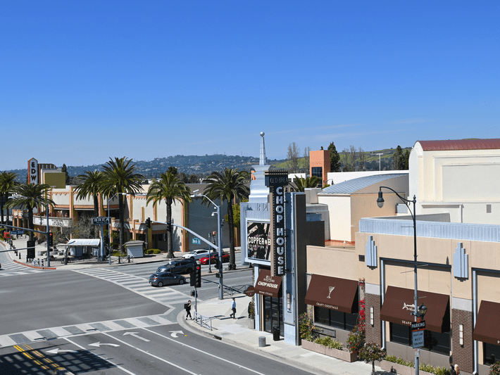 A sunny street scene features palm trees, shops, and restaurants in a modern shopping district. Few people cross the street at a crosswalk, and cars are absent from the roads. Blue sky and distant hills are visible.