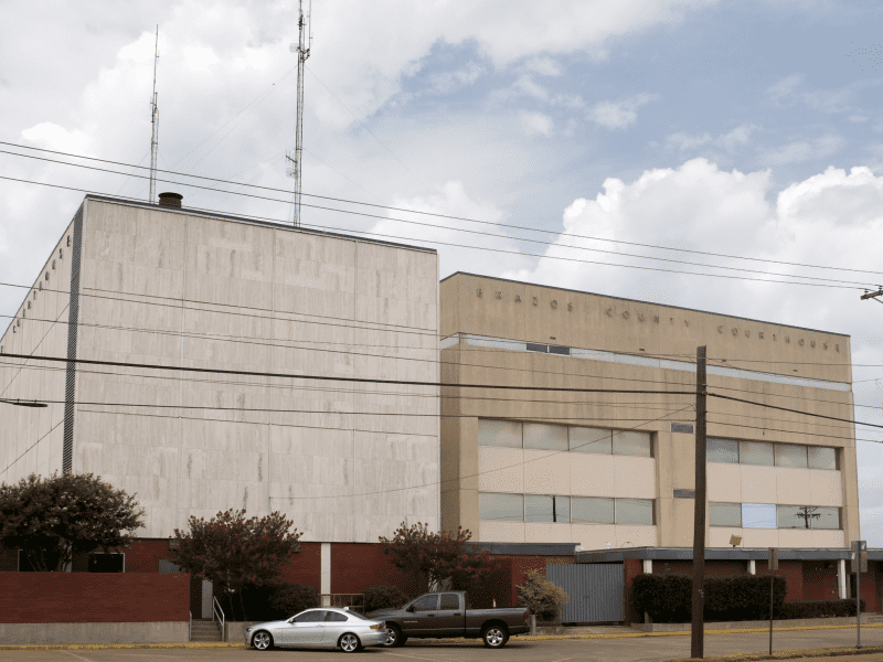 A beige, rectangular courthouse building with minimal windows, tall radio antennas on the roof, parked cars in front, and power lines crossing the scene under a partly cloudy sky.