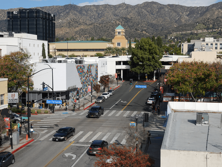 A city street intersection with cars, crosswalks, pedestrians, and buildings, set against a backdrop of tree-covered hills and mountains on a clear day. A blue San Fernando street sign is visible.