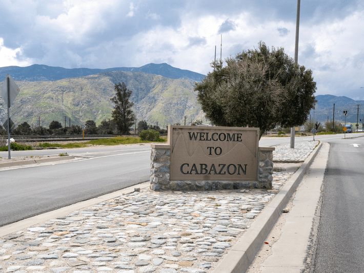 A stone sign reading Welcome to Cabazon stands beside a road with mountains and greenery in the background under a partly cloudy sky.