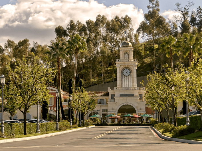 A grand clock tower with an arched entrance stands at the end of a tree-lined road, surrounded by parked cars, green umbrellas, and outdoor seating, with a hillside and partly cloudy sky in the background.