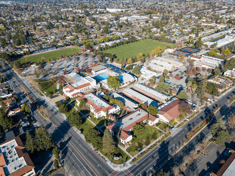 Aerial view of a large school campus with multiple buildings, parking lots, sports fields, and surrounding trees, situated in a suburban neighborhood with houses and streets nearby.