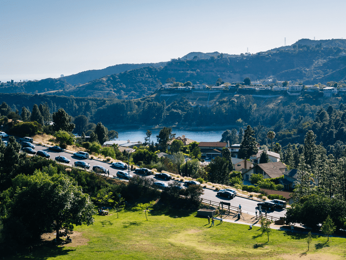 Cars line a road beside a grassy park with scattered trees, as people walk along a path. In the background, hills and houses surround a reservoir under a clear blue sky.