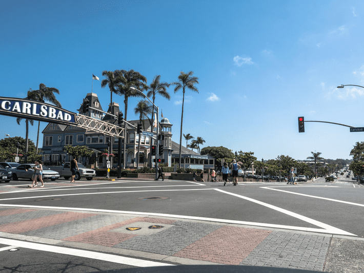 A sunny street scene in Carlsbad, California, with people crossing at a crosswalk, palm trees, a historic building, and a large Carlsbad sign spanning above the road.