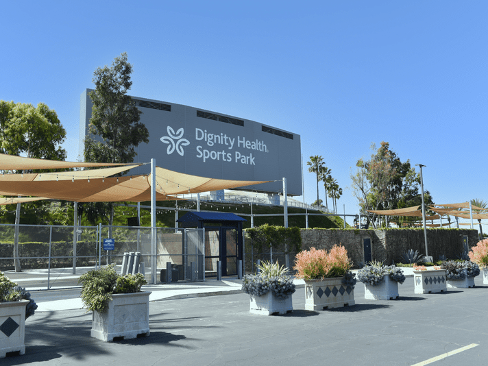 Entrance area of Dignity Health Sports Park, featuring planters with flowers, shade structures, and the stadium building with the venue name and logo visible against a clear blue sky.