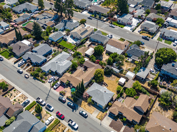 Aerial view of a suburban neighborhood with rows of houses, green lawns, cars parked along the streets, and trees scattered throughout the residential area on a sunny day.