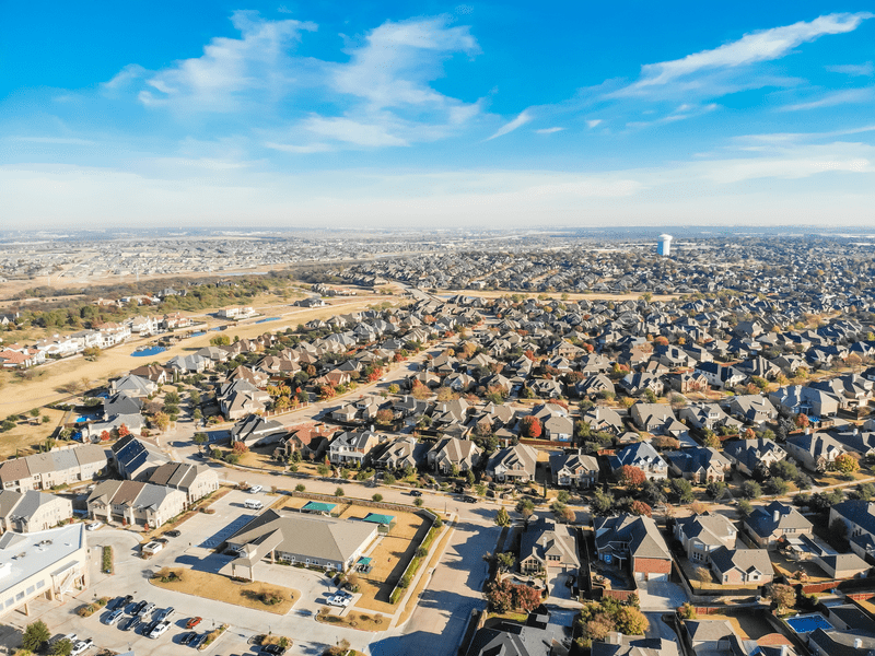 Aerial view of a sprawling suburban neighborhood with rows of houses, green lawns, and wide streets under a bright blue sky with scattered clouds.