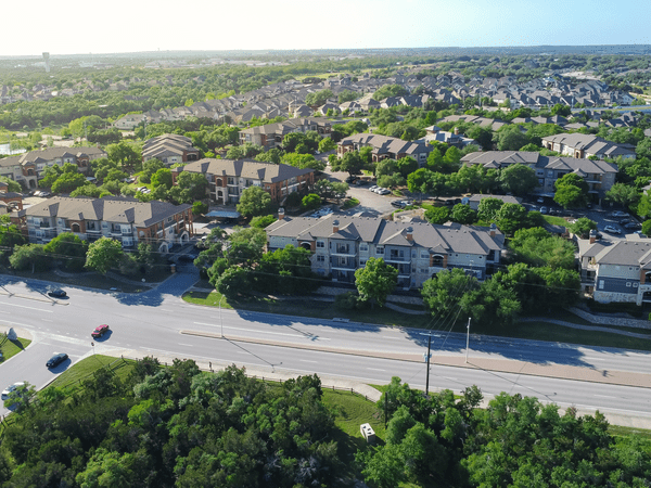 Aerial view of a suburban neighborhood with apartment buildings, green trees, and a main road with a few cars driving. The background shows many houses stretching into the distance under a clear sky.