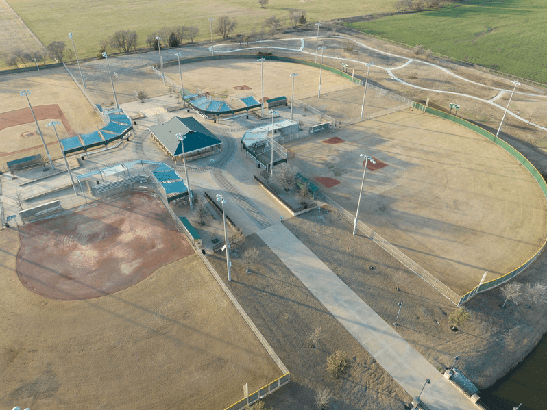 Aerial view of a baseball complex with multiple baseball fields, dugouts, and a central pavilion. The fields have dirt infields and grass outfields, surrounded by fences and light poles, with paths and open grassy areas nearby.