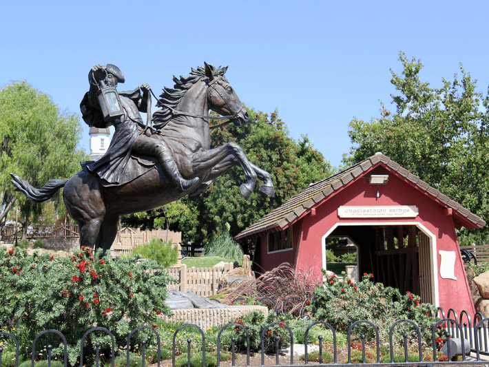 A bronze statue of a rider on a rearing horse stands next to a small red covered bridge surrounded by greenery and flowers under a clear blue sky.
