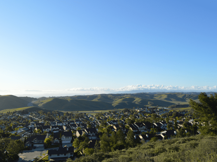A suburban neighborhood sits in the foreground with rows of houses and trees, while rolling green hills and a clear blue sky stretch out in the background.