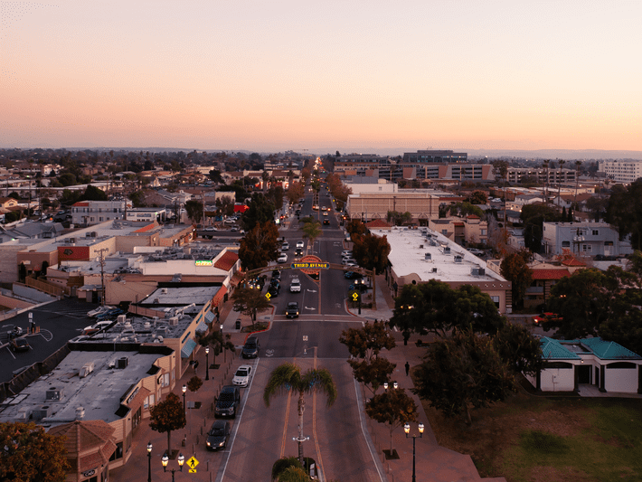 Aerial view of a small city at sunset, showing a main street lined with buildings, cars, and trees, with distant mountains under a clear sky.