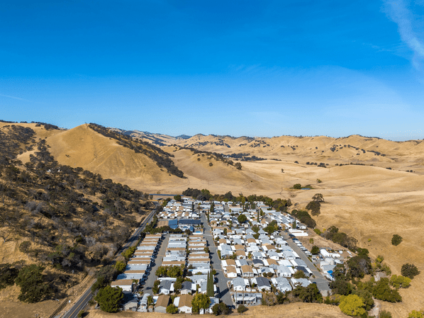 Aerial view of a small residential community with white rooftops, nestled among dry, rolling golden hills under a clear blue sky. Sparse trees and winding roads surround the settlement.