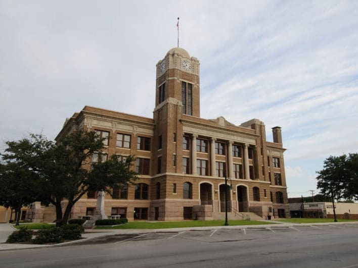 A large, historic brick courthouse with a clock tower and arched windows stands on a corner lot, surrounded by green grass, trees, and an empty street under a cloudy sky.