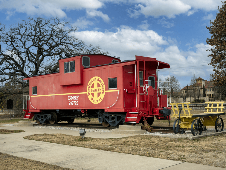 A bright red BNSF Santa Fe Railway caboose is displayed outdoors on a short track section, next to a yellow handcar, with trees and a historic building in the background under a partly cloudy sky.