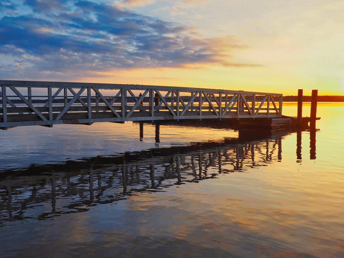 A wooden pier extends over calm water at sunset, reflecting the vibrant colors of the sky—gold, orange, and blue—on the surface. The scene is tranquil and serene.