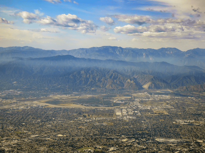 Aerial view of a sprawling city bordered by green hills and distant mountain ranges under a partly cloudy sky. Urban and industrial areas are visible in the foreground.