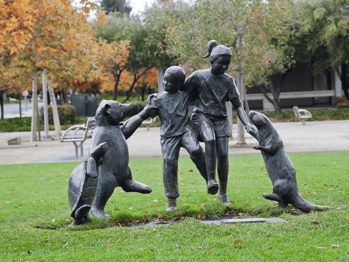 Bronze statue of two children holding hands and playing with a turtle and an otter on a grassy area, with trees and benches in the background.