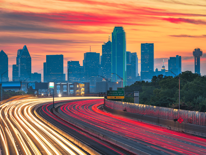 A long-exposure photo of a busy highway with light trails from cars, leading into the Dallas skyline at sunset. The sky glows orange and pink, with skyscrapers illuminated, including one outlined in green lights.