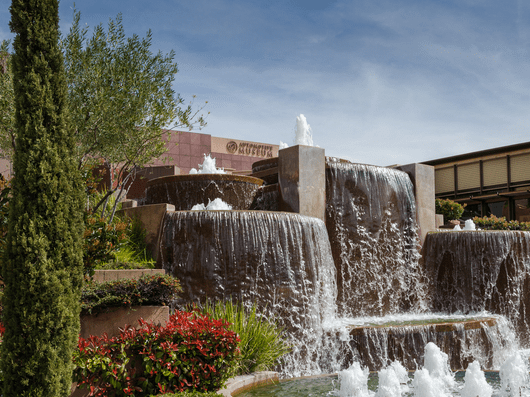 A cascading multi-level water fountain surrounded by lush greenery and red flowers, with a building in the background under a blue sky.