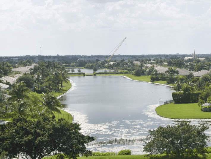 A wide canal or lake lined with palm trees and houses on both sides, with a grassy area in the foreground and a large construction crane visible in the distance under a partly cloudy sky.