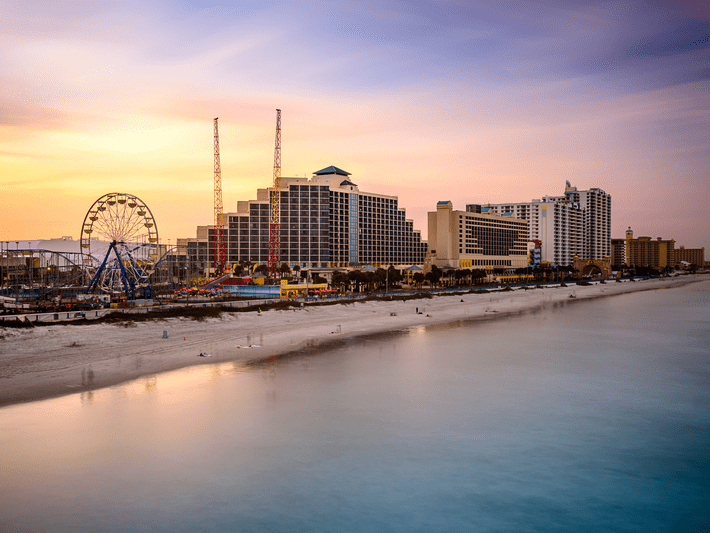 A beachside cityscape at sunset with hotels, an amusement park featuring a Ferris wheel and cranes, and a calm ocean in the foreground. The sky is painted in soft pastel hues.
