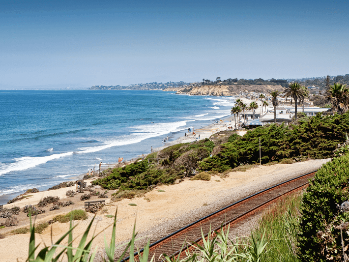 A scenic coastal view with a sandy beach, ocean waves, palm trees, green bushes, and railroad tracks running parallel to the shore under a clear blue sky. People are visible enjoying the beach in the distance.